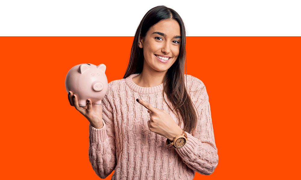 em fundo laranja e de camisa rosa, mulher segura um cofrinho branco de porco com a mão esquerda e com a mão direita aponta para ele.