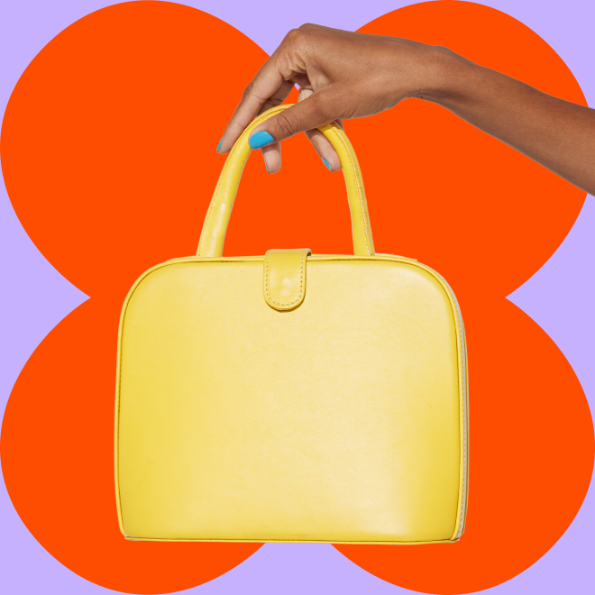 Modelo de perfil olha para a câmera por entre as alças de uma bolsa, que ela segura de lado e apoia no ombro.