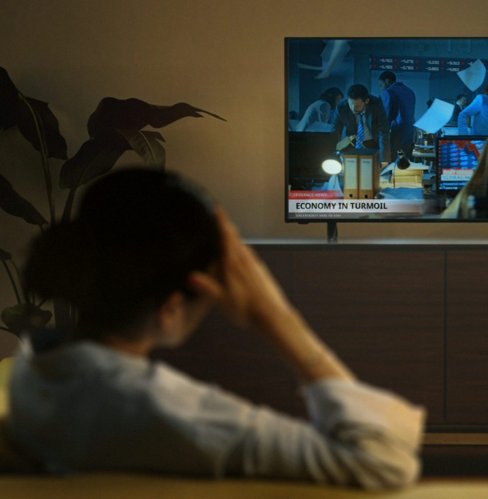 A retail gold investor sits in her living room watching TV. On-screen, we see a news channel reporting a financial crisis.