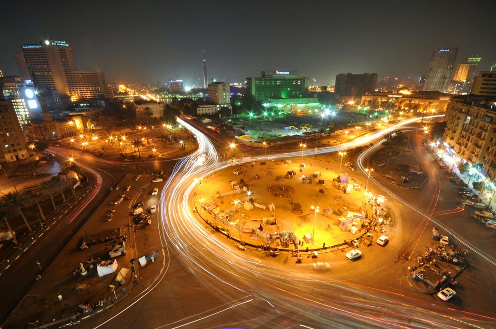 A shot of a large roundabout in the city at dusk–as if taken from a tall building—illuminated in gold light by car headlamps