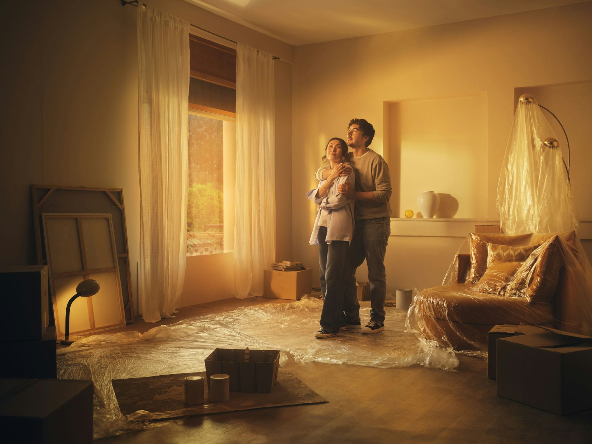 A couple stands in the living room of their new house. From the protective sheets covering the furniture, they are decorating