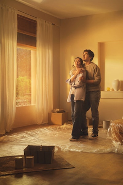 A couple stands in the living room of their new house. From the protective sheets covering the furniture, they are decorating