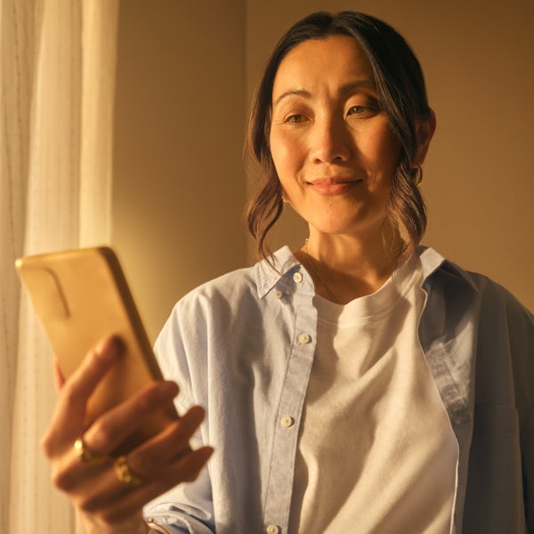 A woman holding her phone and investing in gold online. She is smiling and has a golden glow. There's a window to her right.