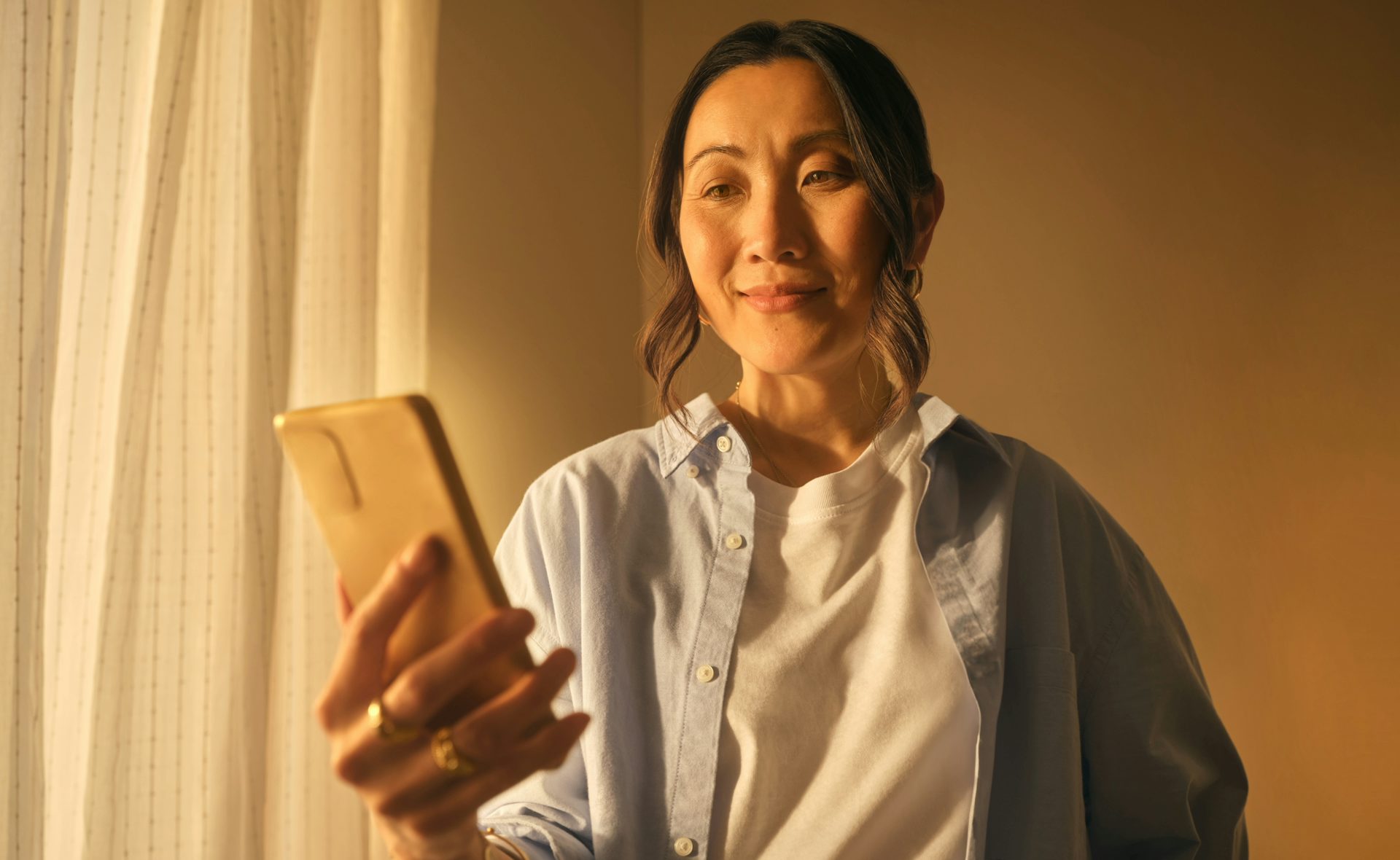 A woman holding her phone and investing in gold online. She is smiling and has a golden glow. There's a window to her right.