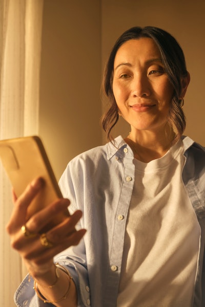 A woman, aged 30 to 45 years, is holding her phone, looking at the screen and smiling. She is learning how to own gold online