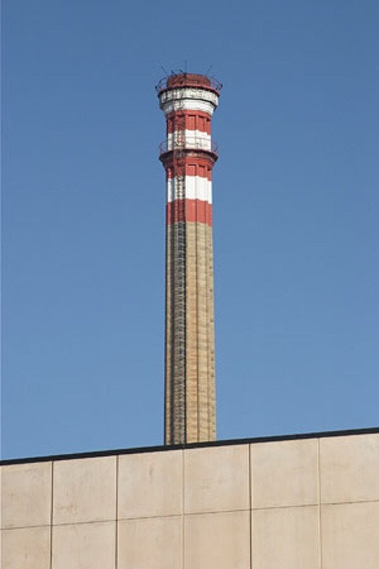 Ciminiera alta e strutturata con strisce rosse e bianche, emergente dietro un edificio industriale con cielo blu chiaro sullo sfondo.