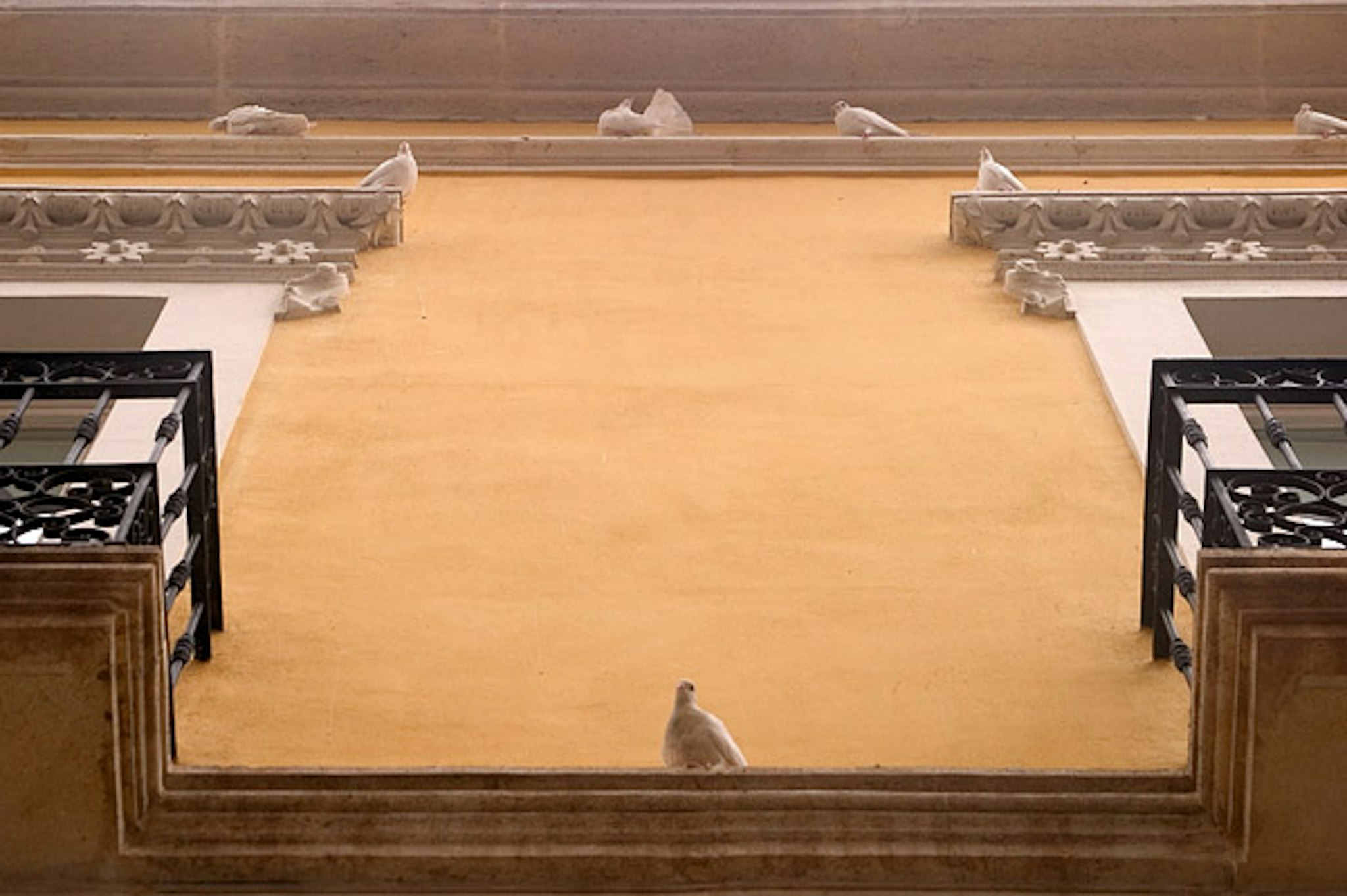 Una vista dall'alto di un cortile interiore con una balconata dotata di guardrails in legno e delle colonne decorate in pietra bianca con strapiature. Il piano è provvisto di parcheggi a lato senza nessuna persona presente.