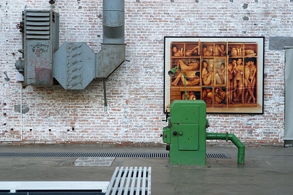 Edificio industriale con muro di mattoni come sfondo. Una stampiera a vapore vecchia sta accanto a una grande muralesca di figure nude. C'è anche un pompino verde in fronte alla muralesca, parzialmente coperta da esso. L'ambiente ha un'aria artisticamente industriale.