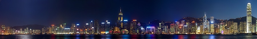 The photo shows an urban night scene with a city skyline in the background, illuminated by artificial lights. There are silhouettes of buildings against a dark sky, suggesting it is evening or nighttime. In the foreground, there is a body of water that reflects some of the light from the buildings. The style of the image is a straightforward photograph without any filters or artistic effects.