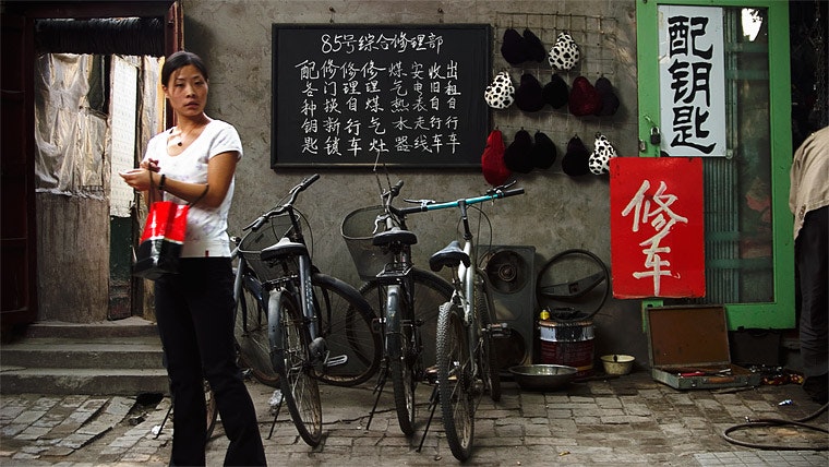 Un individuo sta vicino a una cartellina in cinese, tiene un sacchetto rosso e ci sono dei biciclette parcheggiate vicino.