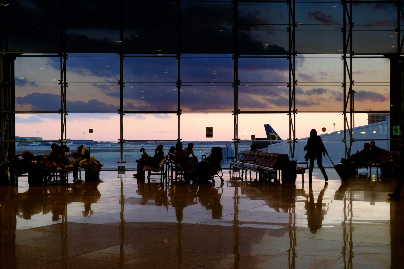 In un terminale aeroportuale, diverse persone sedute e in piedi sono silhuettate contro una grande finestra di vetro, attraverso cui è visibile un aereo sulla pista e un cielo al tramonto, creando un'atmosfera di viaggio e attesa.