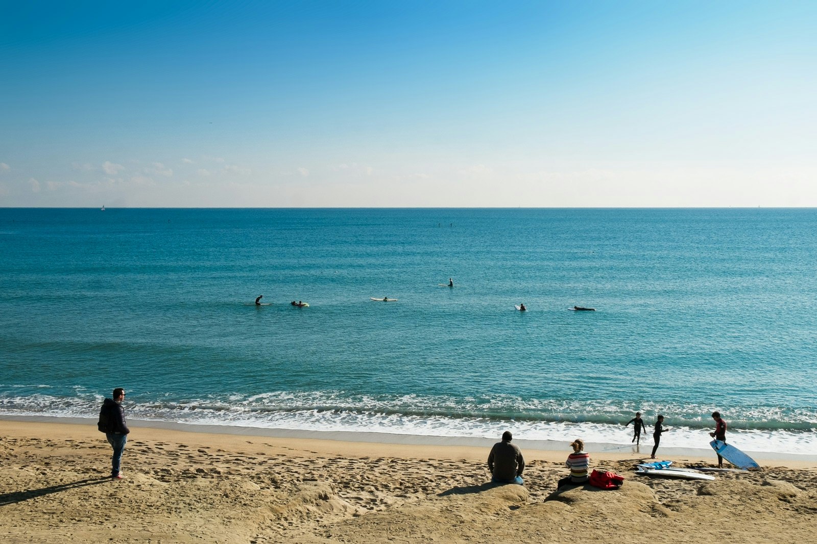 A vibrant beach scene with blue water, people playing in the sand, and surfers enjoying the waves.