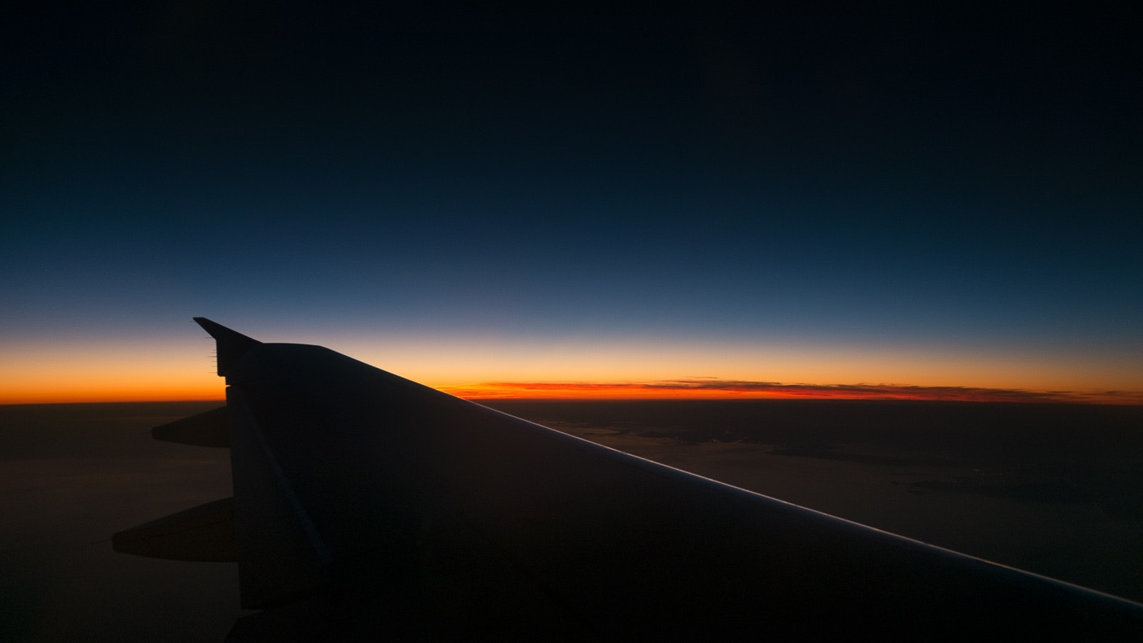 This is an image of a sunset with airplane windows, possibly taken from the window of an airplane. The sky transitions through shades of pink, orange, and blue. There appears to be a small section of the wing visible at the bottom of the photo.