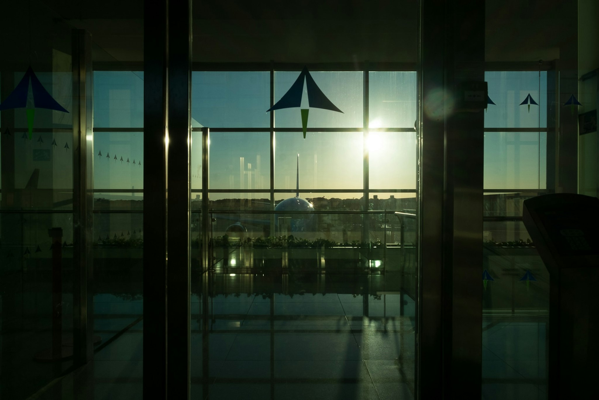 Vista di una città al tramonto attraverso una finestra di vetro in un edificio, con il sole che brilla intensamente all'orizzonte e un'architettura moderna visibile in lontananza.