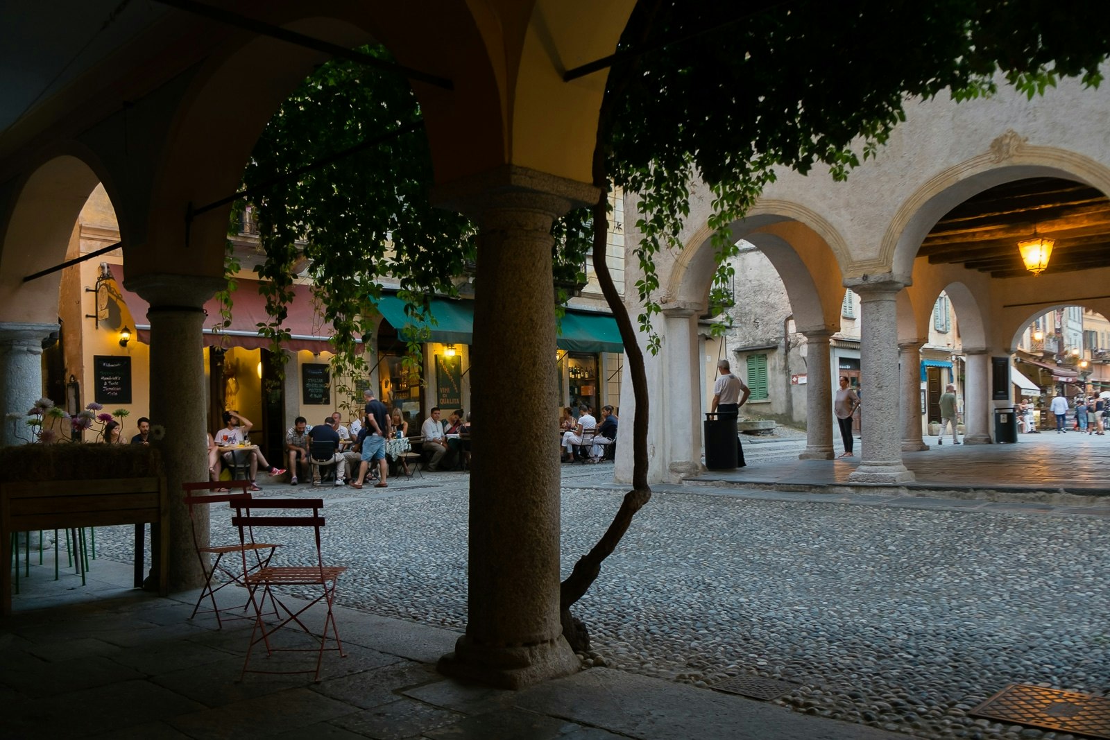 Una serata tranquilla in una piazza storica con portici e tavolini all'aperto, illuminata da luci calde, mentre persone si godono una cena al ristorante; vi sono archi in pietra e decorazioni in verde che aggiungono fascino al paesaggio urbano mixando elementi architettonici antichi.