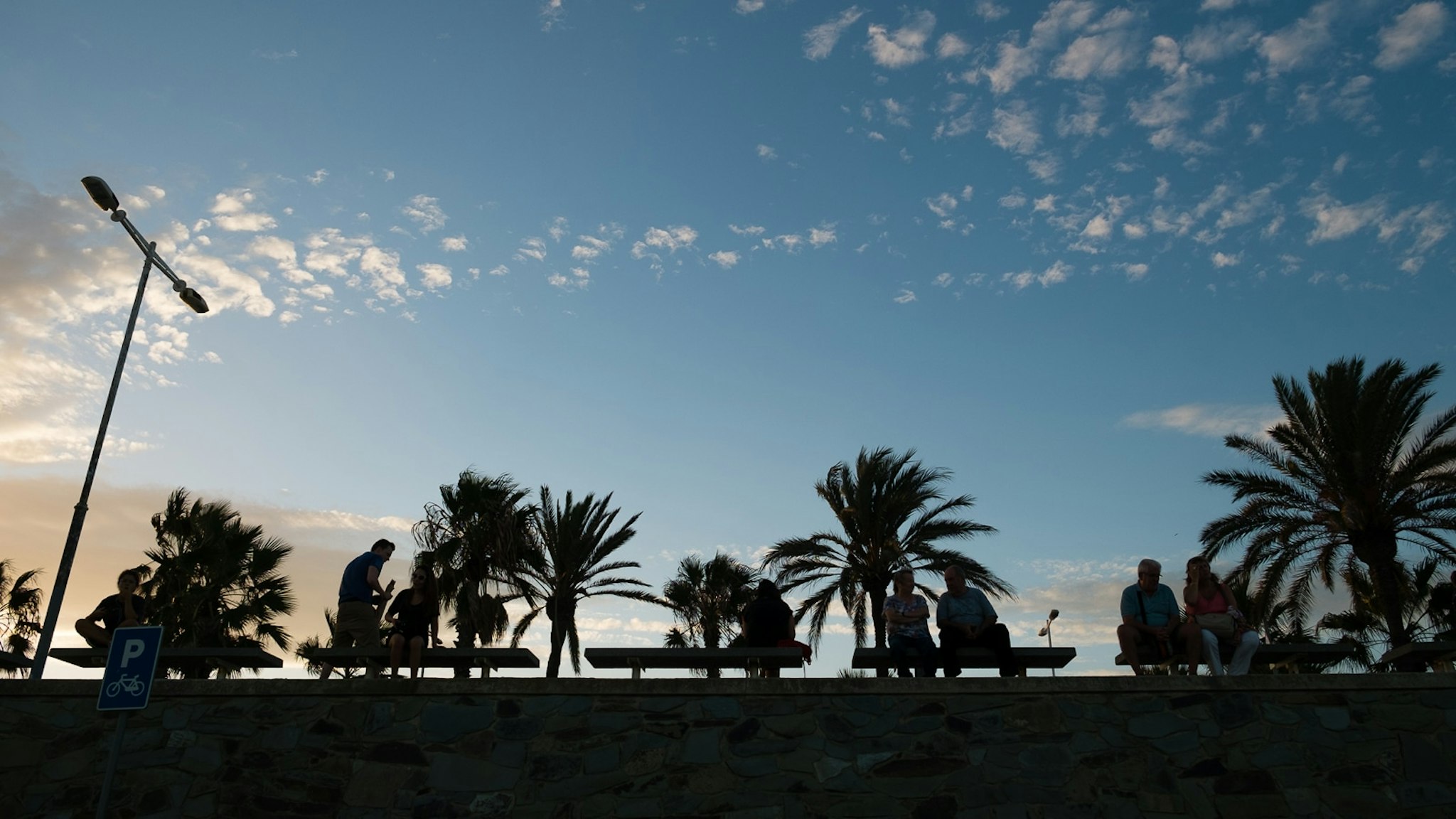 Una vista panoramica di una spiaggia durante il tramonto con persone che godono dell'ora notturna. Il cielo è dipinto di colori caldi riflessi sull'acqua. Palme d'albero si agitano leggermente sul fondo del tramonto.