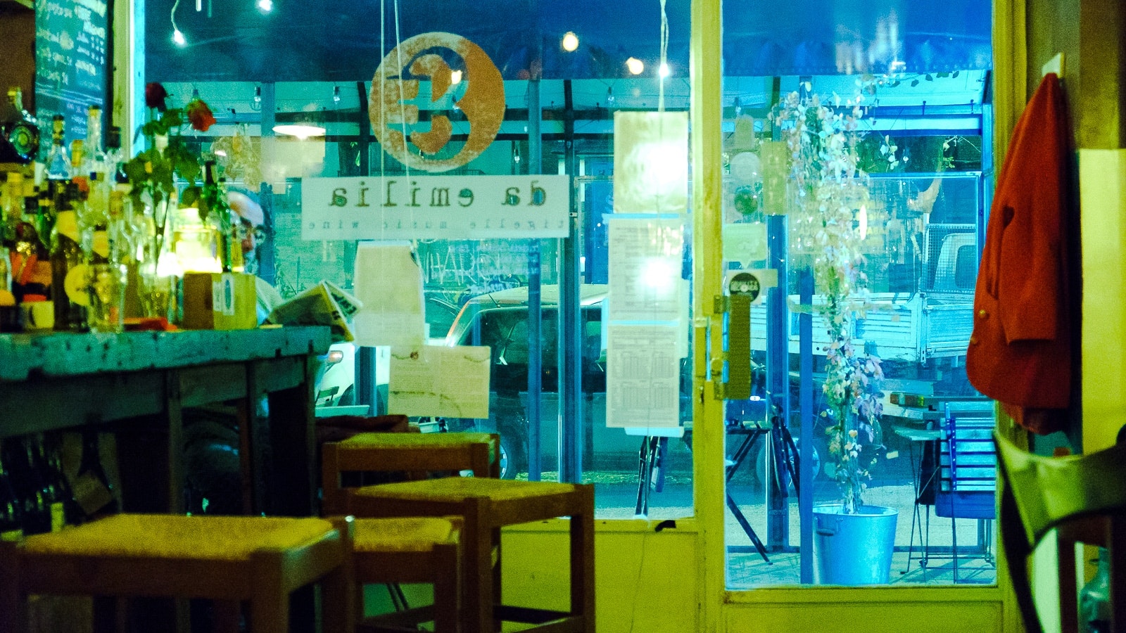 Una ristorante intimo accogliente, vista attraverso una finestra durante la notte. L'interno ha una illuminazione scura e è dotato di tavoli e sedie, mentre un segnale all'esterno indica che è aperto per i clienti.