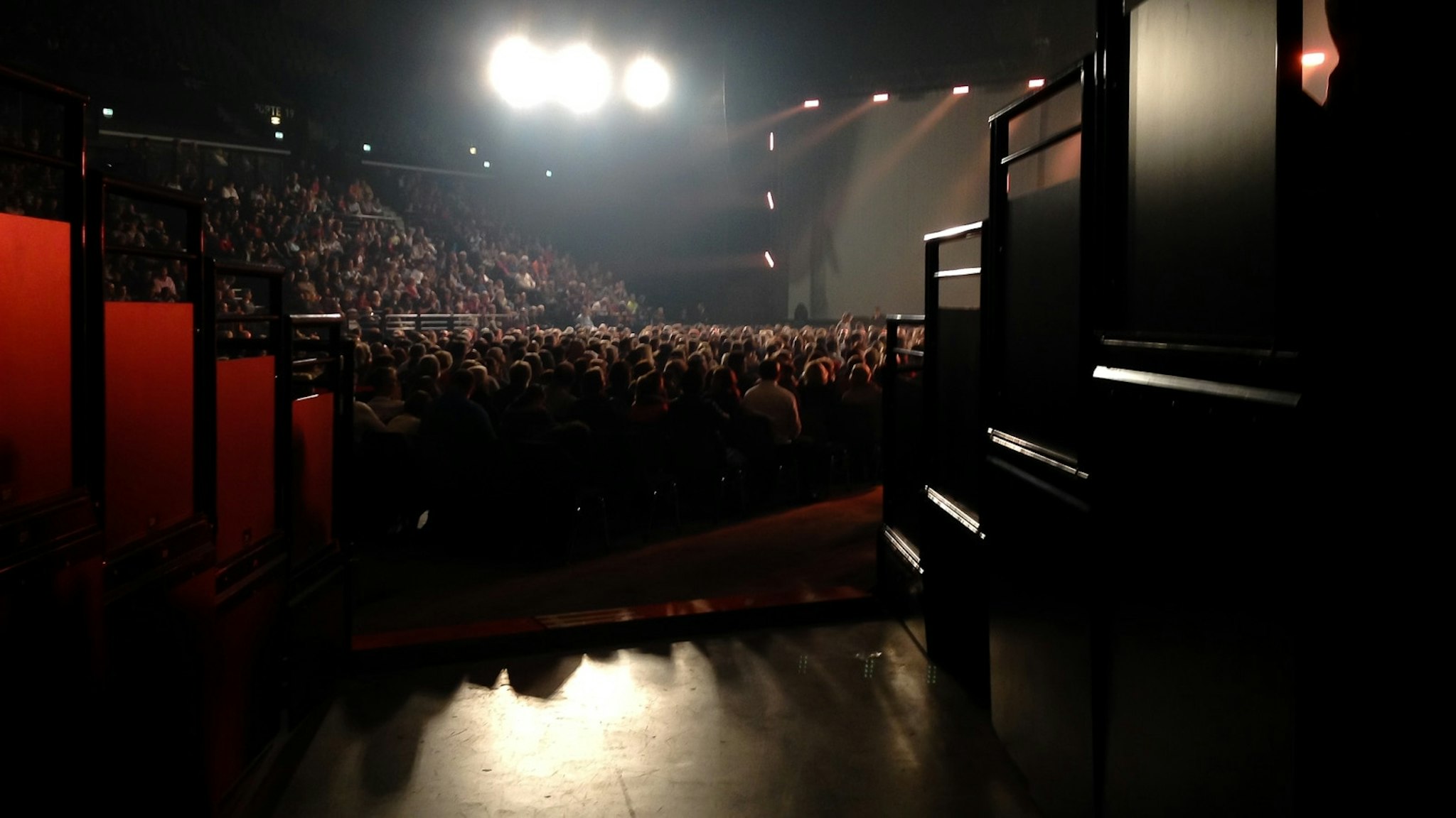 Anfiteatro con sfondo oscuro, mostra un artista sul palco di fronte a un pubblico, visto dalla terza fila.