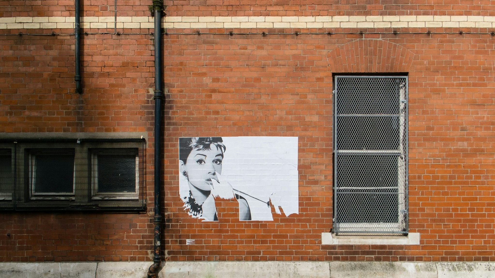 Brick wall con affiche di una persona, vicino ad un finestrato chiuso. L'affiche pare parzialmente coprito da un foglio in frantumi.