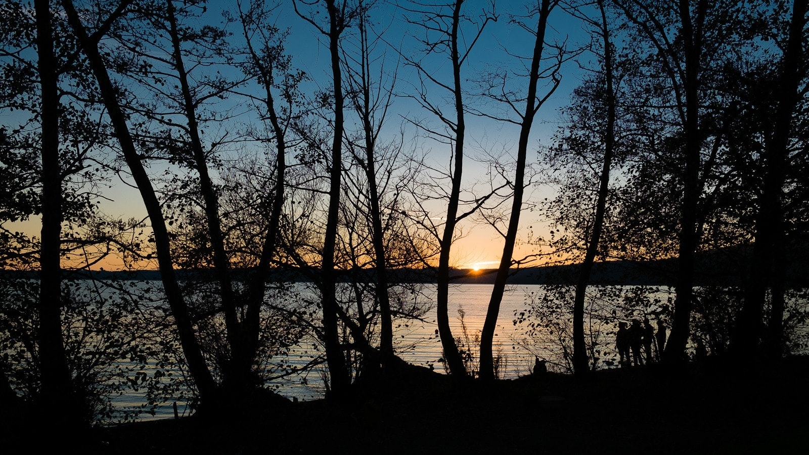A tranquil lakeside scene during sunset, with trees and reflections on the water's surface. The sky transitions from blue to orange with the setting sun.