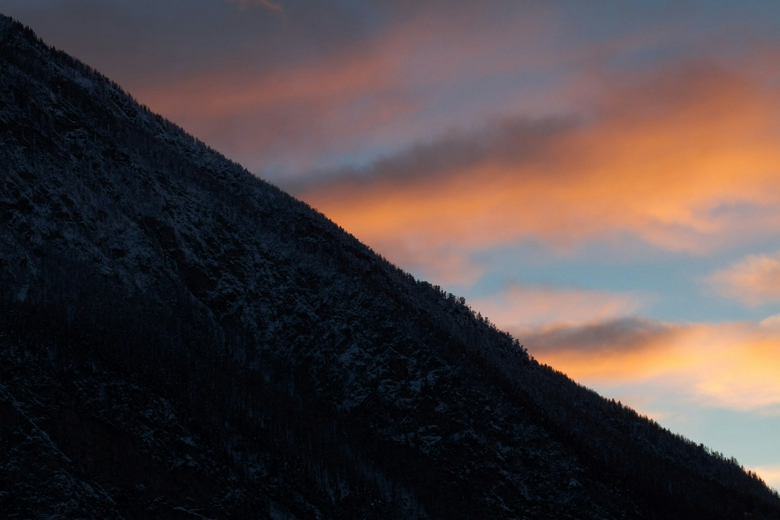 A stunning mountain sunset with the majestic peak silhouetted against the vibrant sky, which transitions from a brilliant sunset to the deep blues of dusk. The foreground features a rugged landscape with patches of snow and rocky outcrops under the warm glow of the setting sun.