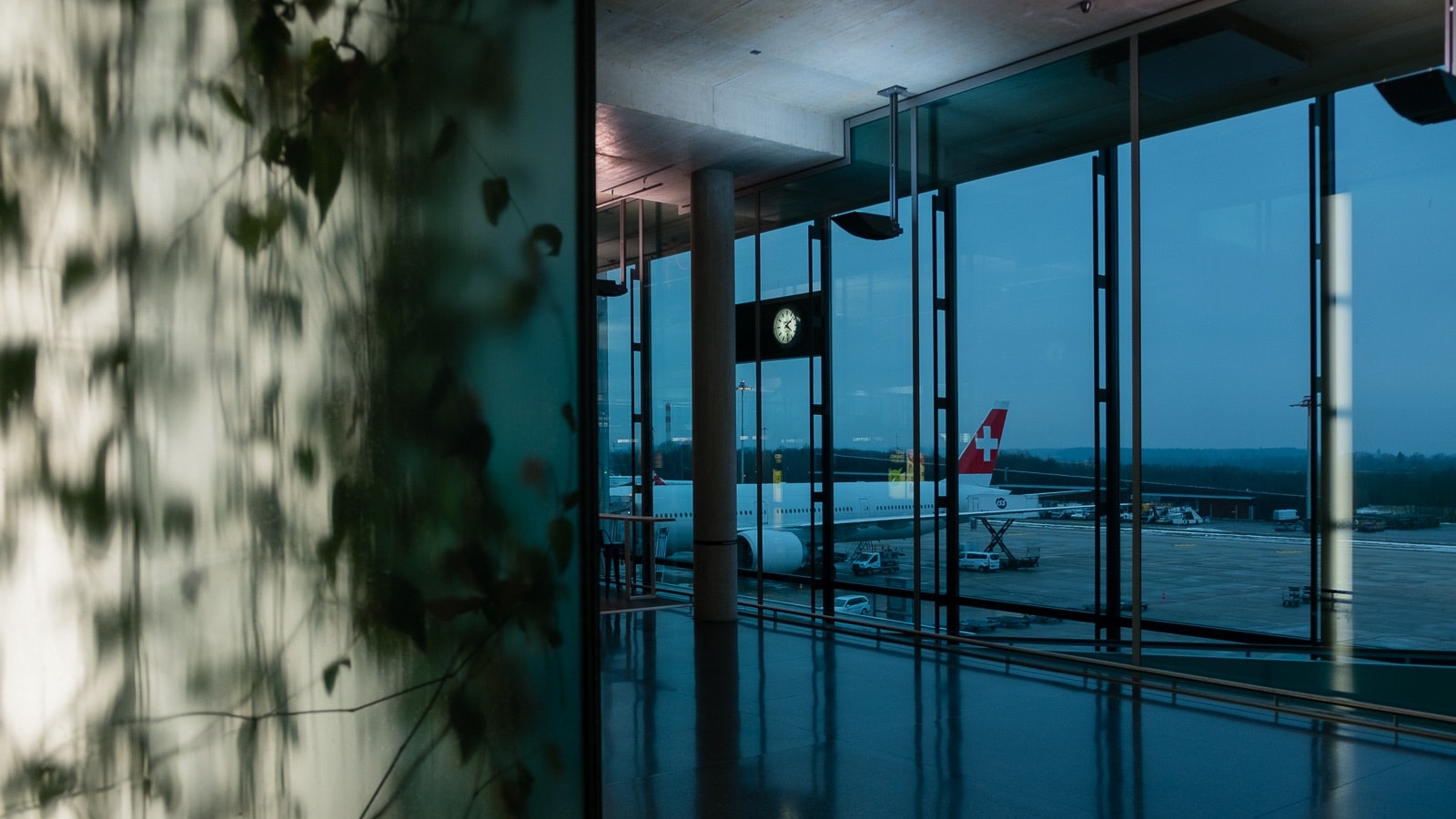 Una foto che rappresenta una stazione di aeroporto notturna, vista dalle finestre della sala d'attesa. Mostra l'esterno del building, un aeroplano sul pavimento e la riflessione sullo vetro che suggerisce un ambiente tranquillo dentro.