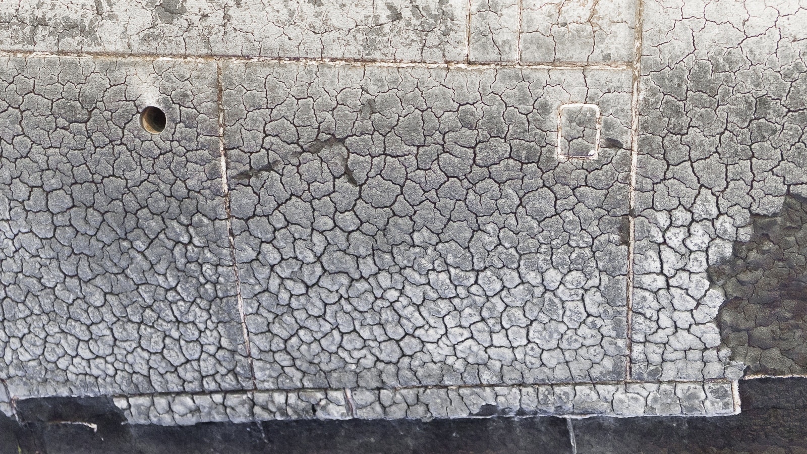 La foto mostra il sottofondo di un edificio o struttura con una pattern di materiali neri sulla superficie, che sembra essere in discontinuità o danno. Inoltre, ci sono tracce con rotondità e testi, che potrebbero indicare un deterioramento o il segnale di una fase di lavoro in corso.