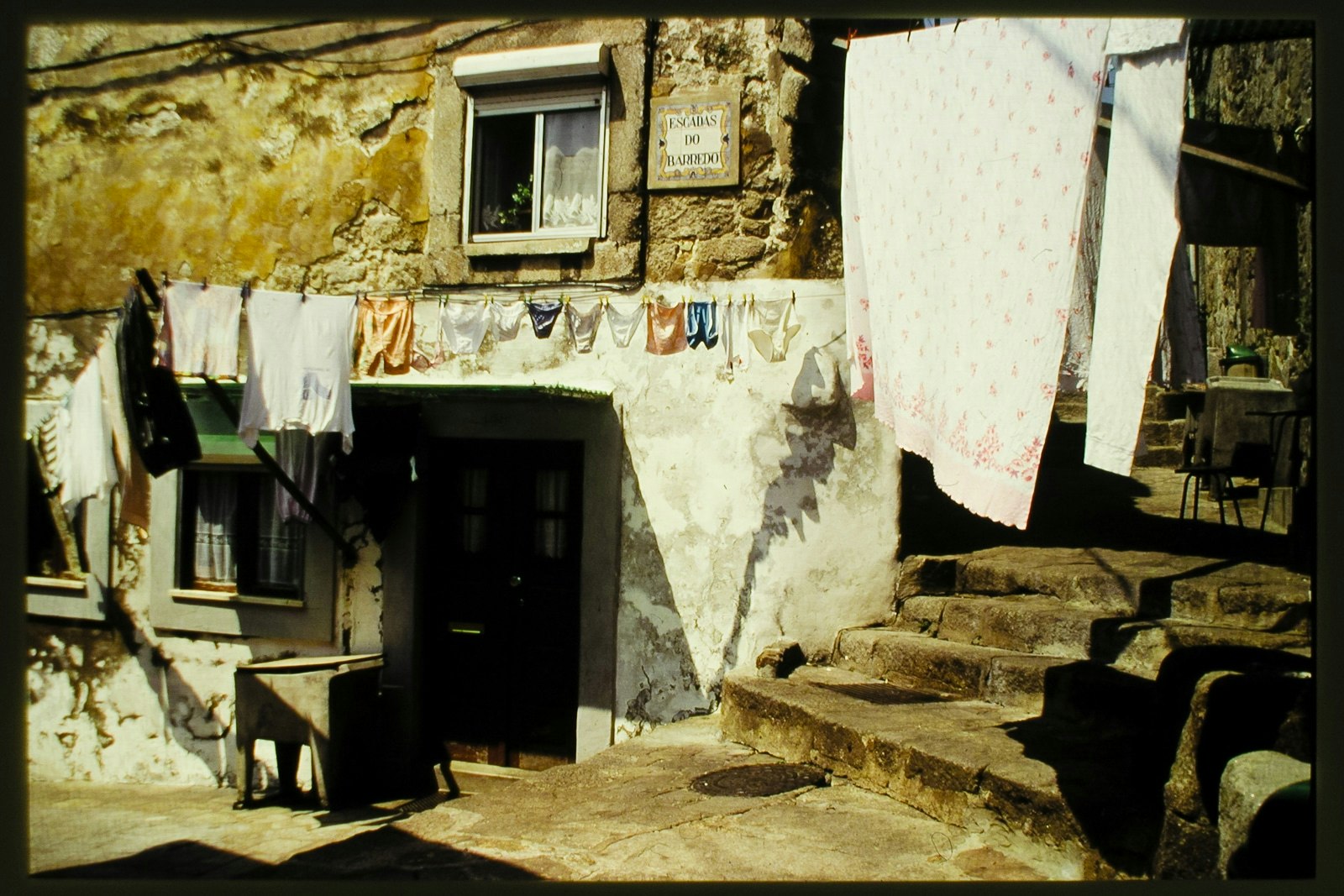 A view of a traditional European laundry area, featuring hanging clothes on a line in front of a building with a sign that's partially obscured, steps leading up to the laundry, and old-fashioned clothes hanging out.