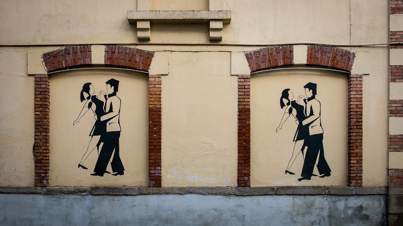 Two identical stencil art paintings on a beige brick wall depict a couple dancing closely, with the vintage urban setting suggesting a sense of romance and nostalgia.