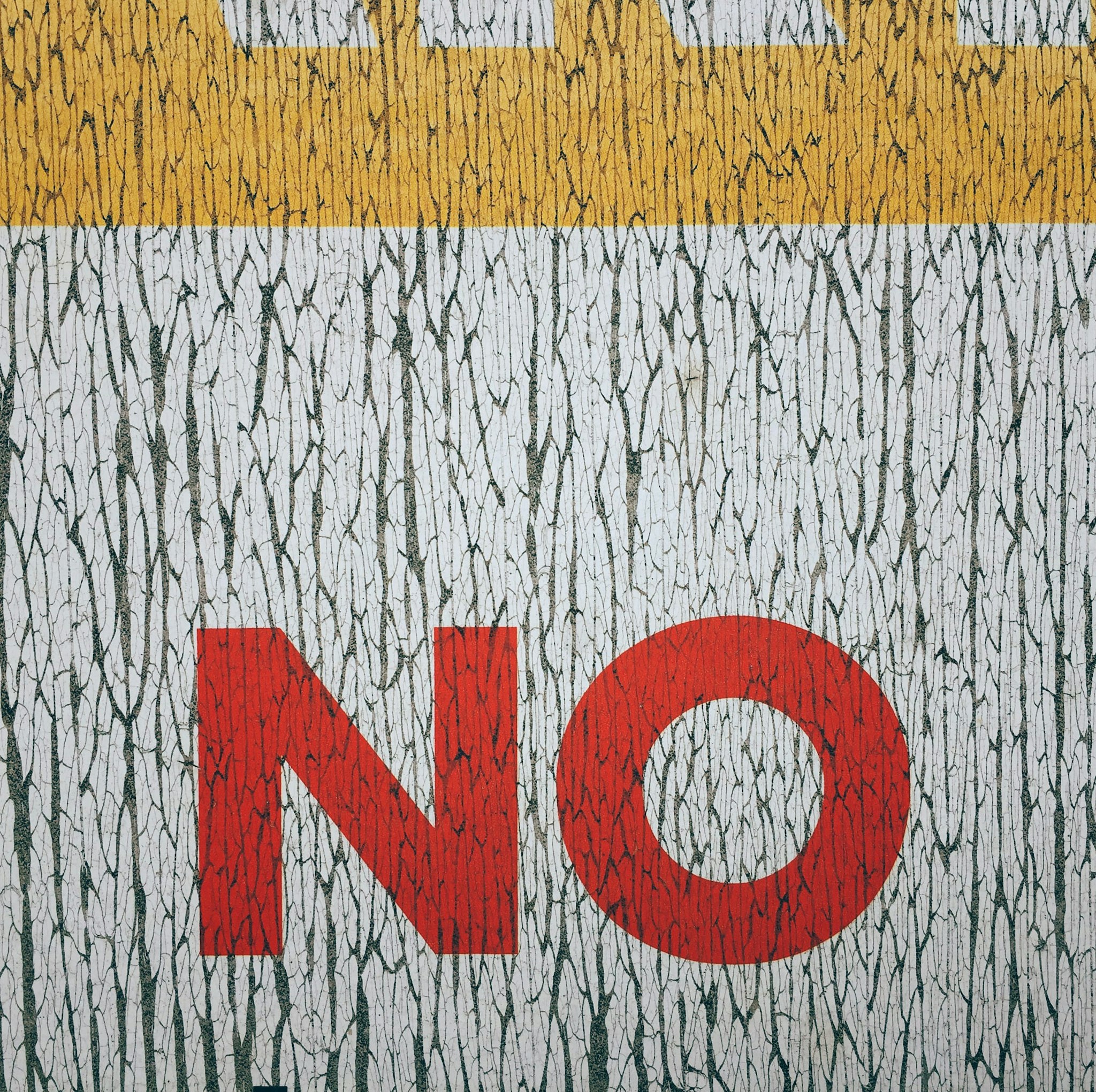 A weathered and cracked painted surface displays the large capitalized word "NO" in bold red letters on a textured white and yellow background.