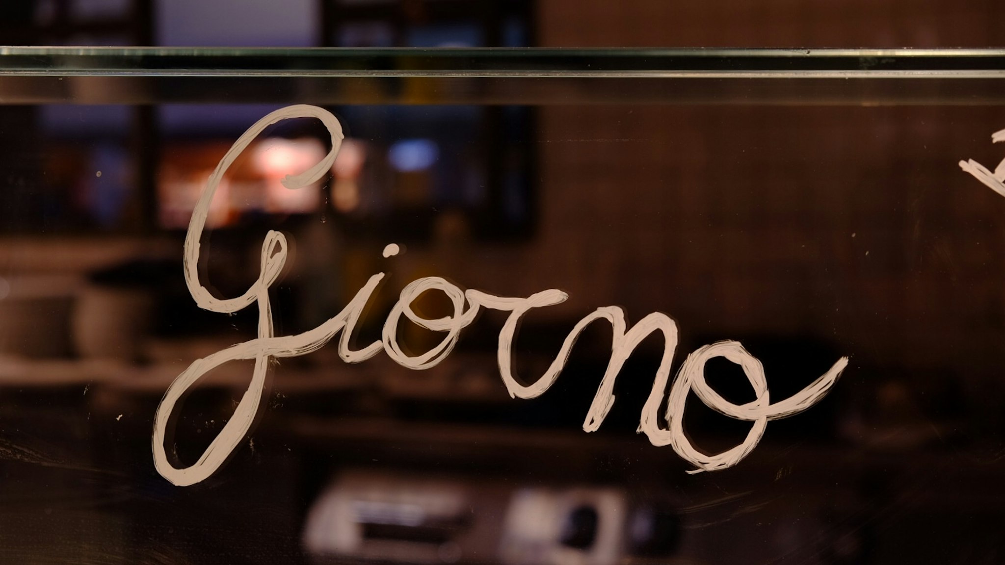 A warmly lit café interior features the word "Giorno" artistically handwritten on a glass panel, with a softly blurred background displaying shelves and kitchenware.