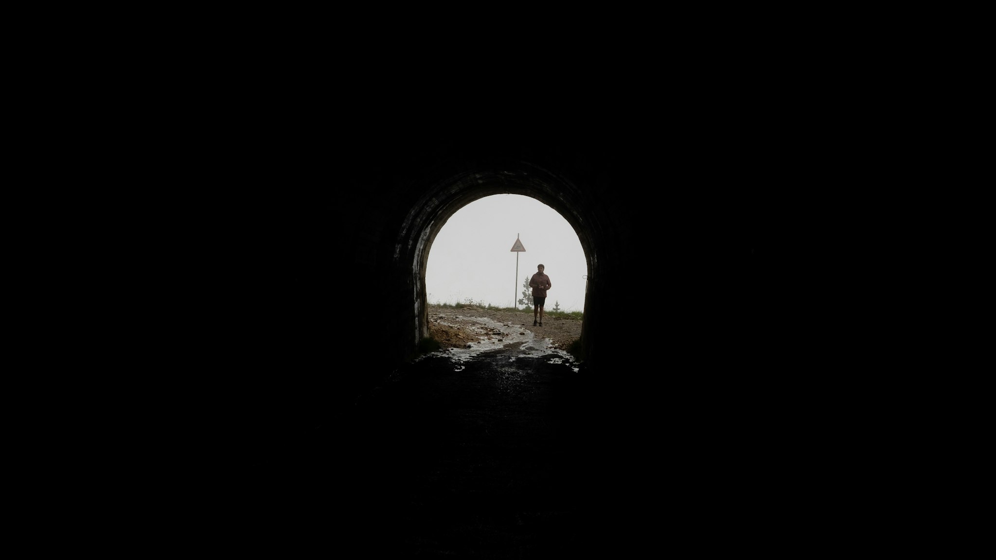 A silhouetted figure runs towards the light at the end of a dark tunnel, highlighting the contrast between shadows and the bright outdoor scene beyond.