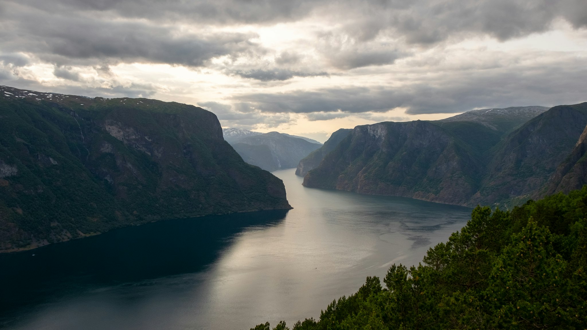 A breathtaking view of a deep fjord surrounded by towering, lush green hills under a dramatic, cloudy sky, reflecting the calm, dark water below.