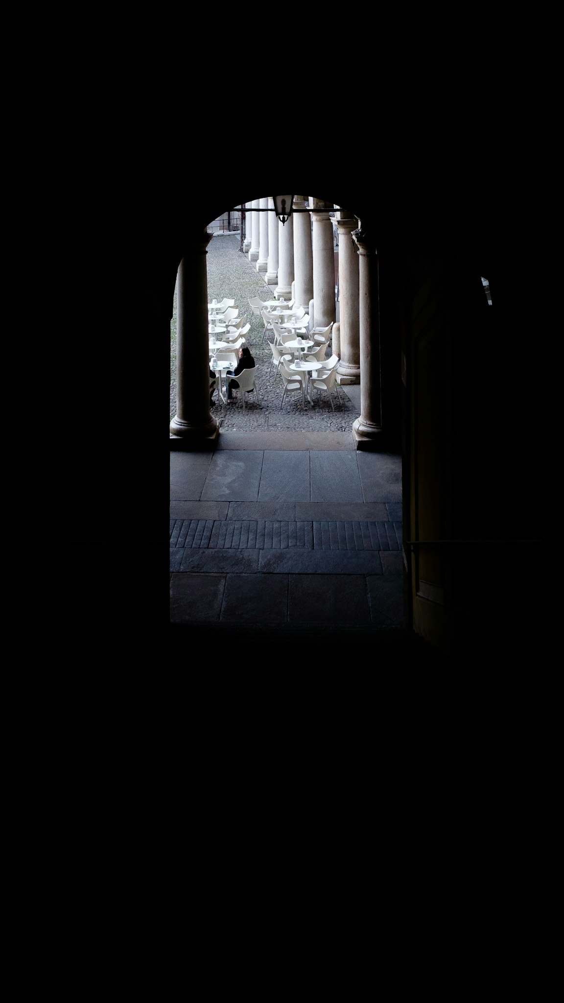 Un arco scuro si apre su una scena luminosa di un caffè all'aperto con tavoli e sedie bianche, delimitato da colonne eleganti.
