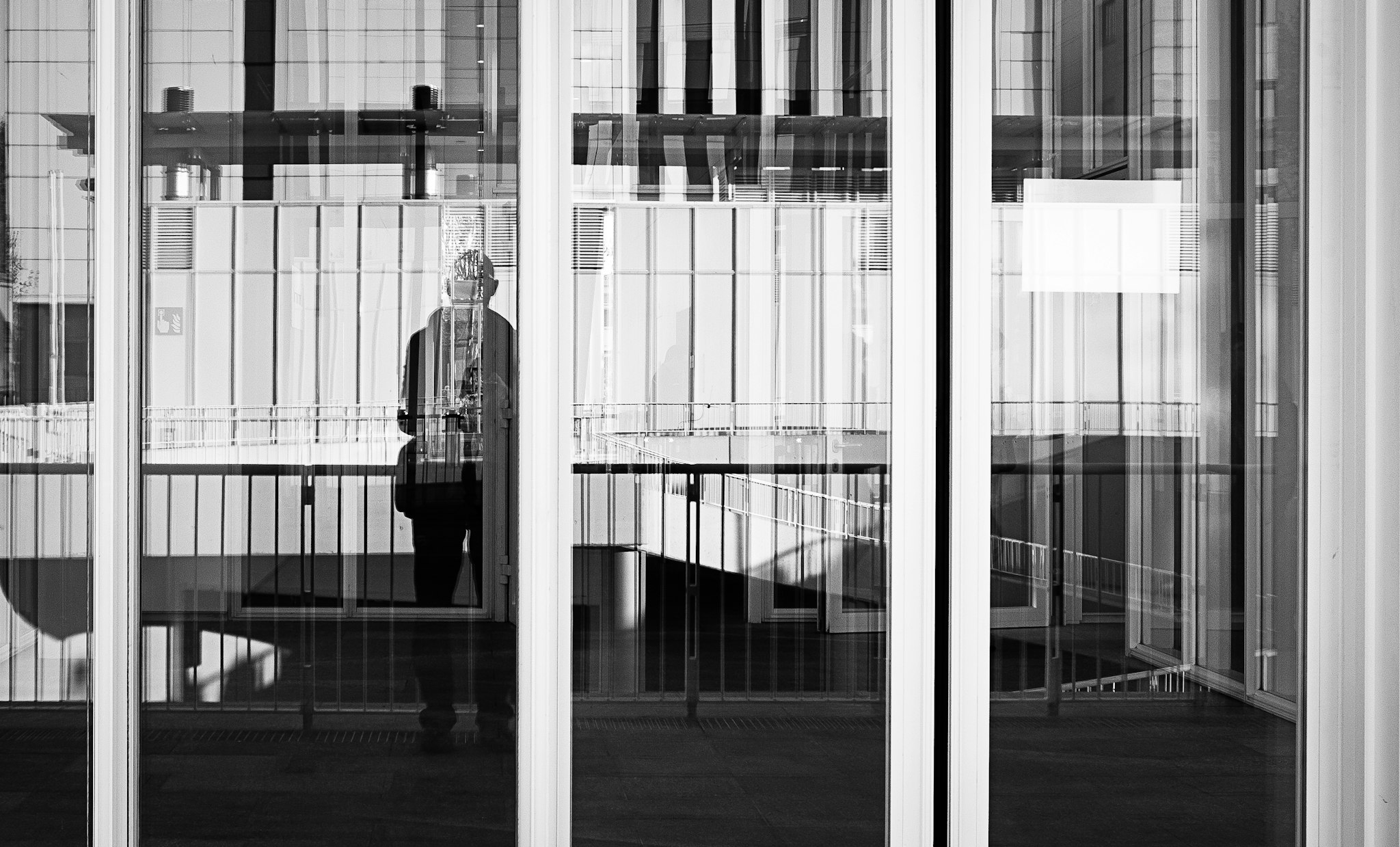 Un uomo sta di fronte a una finestra di un edificio. La riflessione di un uomo è visibile nel vetro della finestra. Alcune finestre sono visibili sulla facciata degli edifici nell'ambiente.