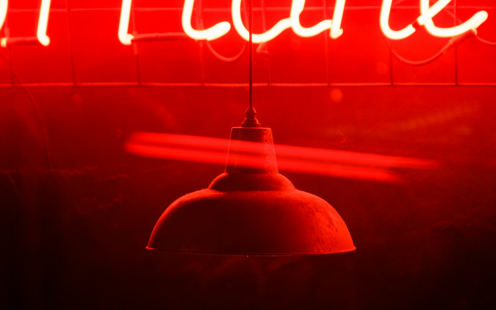 Una lampada a sospensione rustica è illuminata da un'insegna al neon rossa, creando un'atmosfera calda e vibrante in un ambiente oscuro.