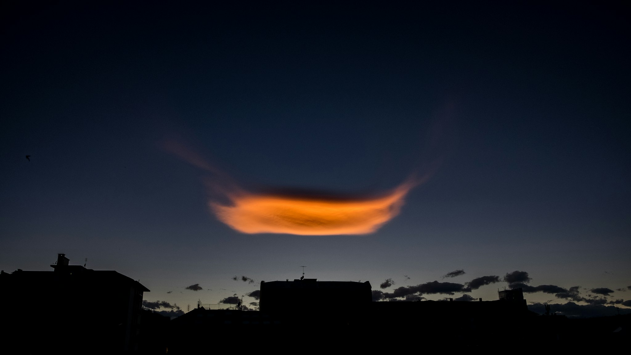 A dramatic sunset sky with a striking orange lenticular cloud floating above silhouetted buildings, creating a captivating and mysterious atmosphere.