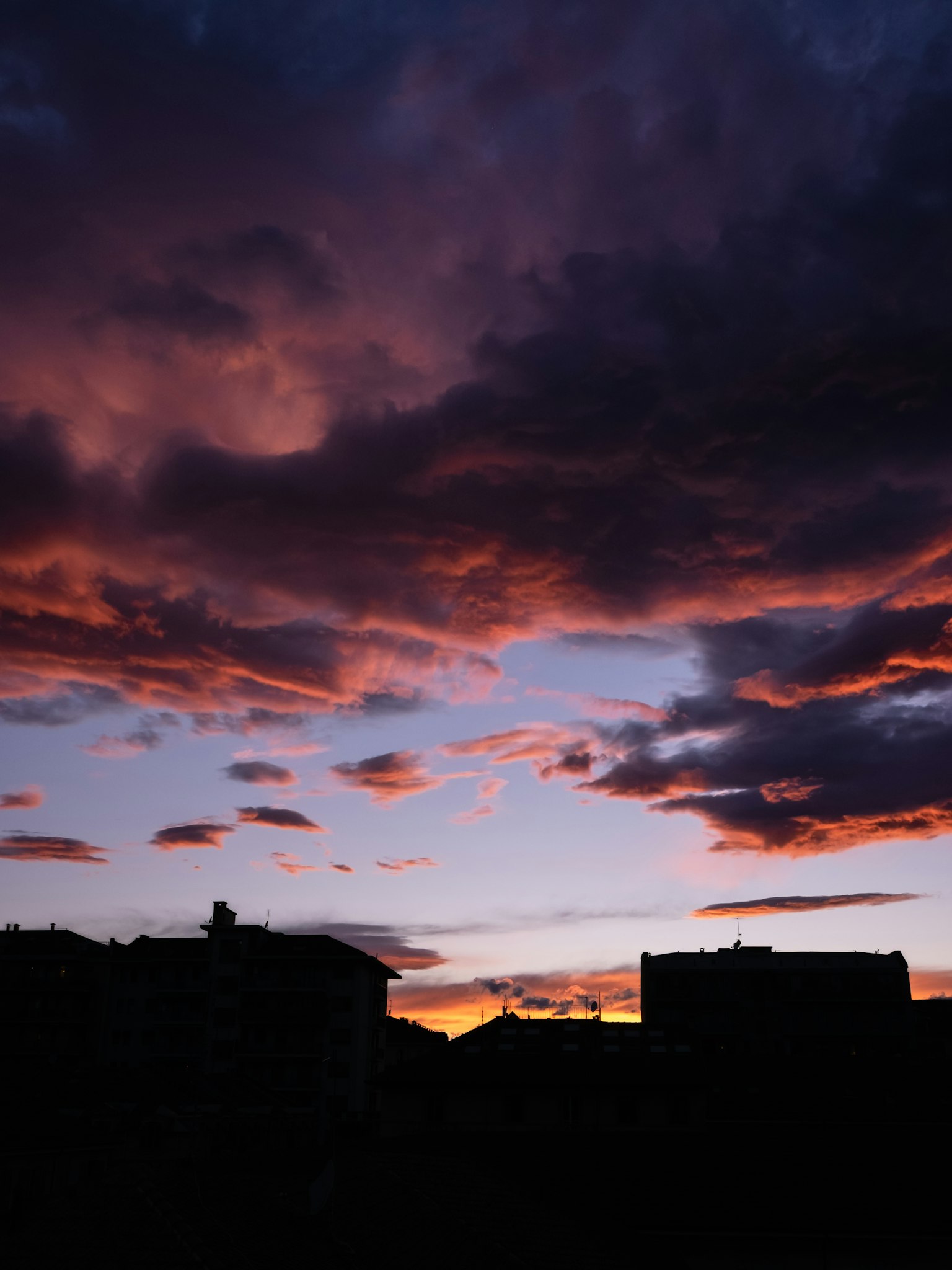A dramatic sunset over a cityscape with silhouetted buildings, featuring vibrant hues of purple, pink, and orange clouds against a deepening blue sky.