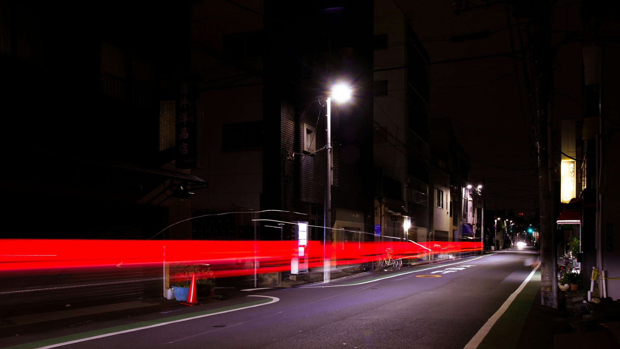 A blurred nighttime shot of a car moving at high speed, with red and white lights from the vehicle streaking through a city street scene, including buildings, traffic lights, and signs in both English and Italian.