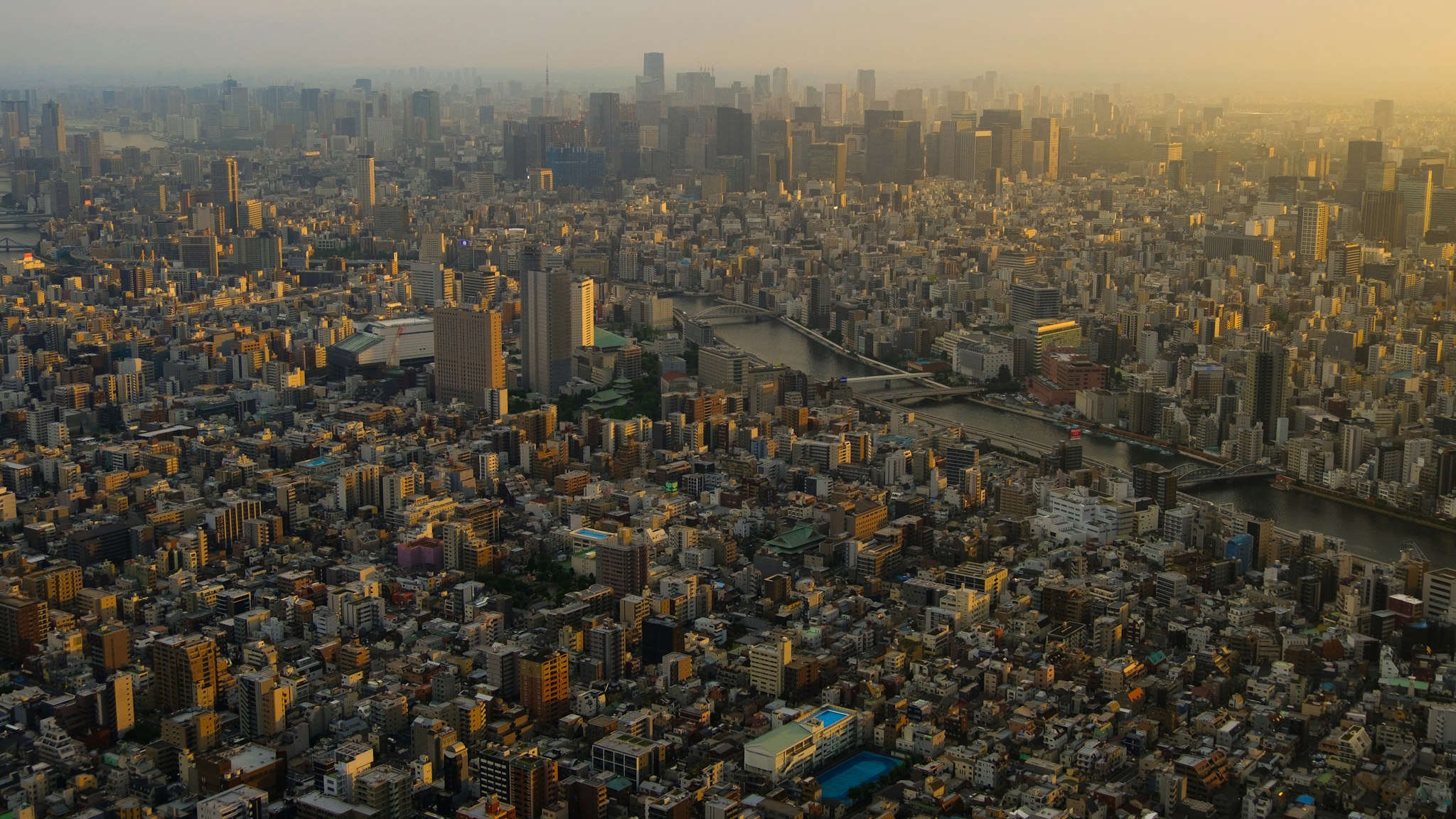 Una veduta panoramica di una metropoli al tramonto con dense aree di edifici e grattacieli, attraversata da un fiume che riflette la luce dorata del sole calante; la città è immersa in un'atmosfera serena e luminescente.
