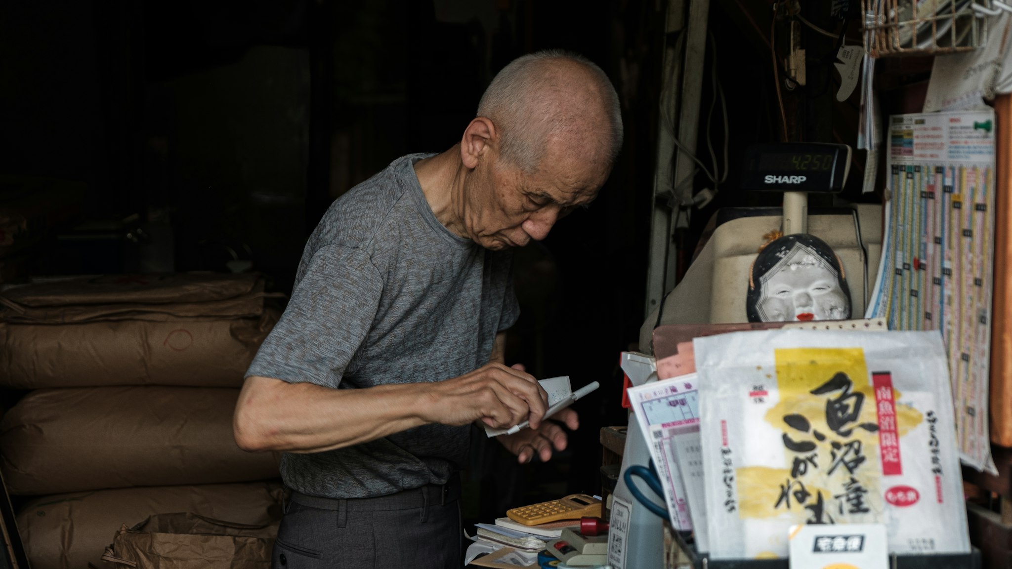 Old man standing at a market stall, reading a label on a product with his right hand.