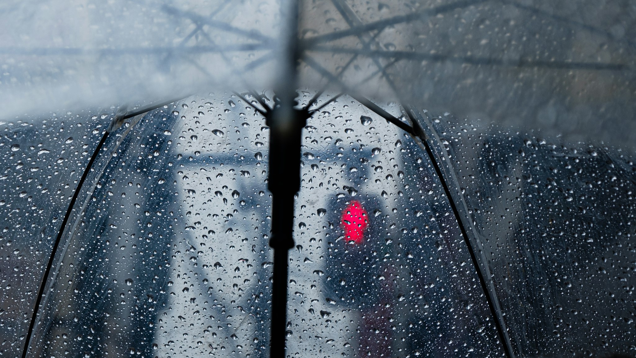 A close-up view of a transparent umbrella covered in raindrops, with a blurred cityscape and a red traffic light in the background, capturing a rainy day in the city.