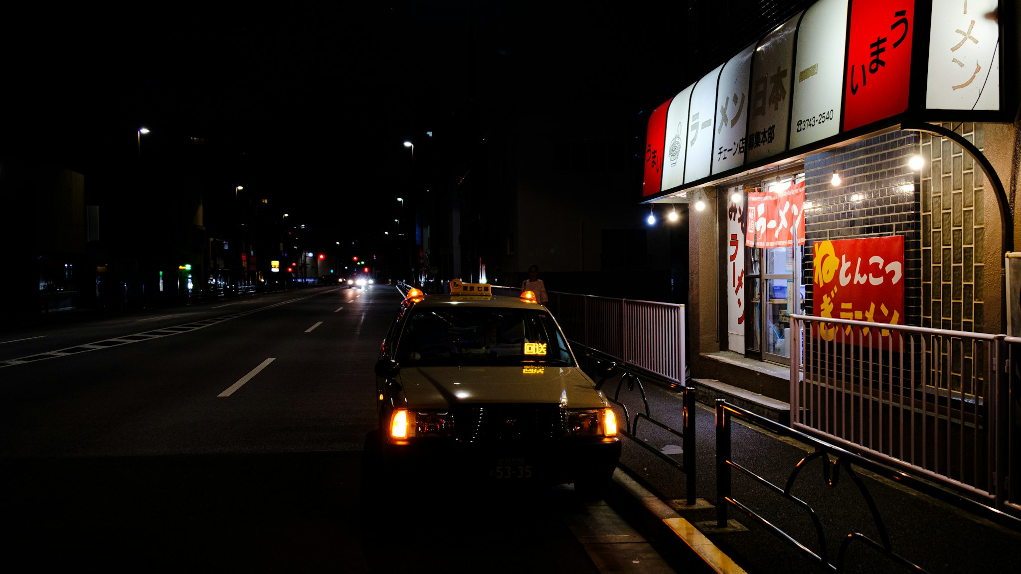A nighttime view of an urban street scene with a lit business sign and dark vehicles on the road, showcasing city life after dark.