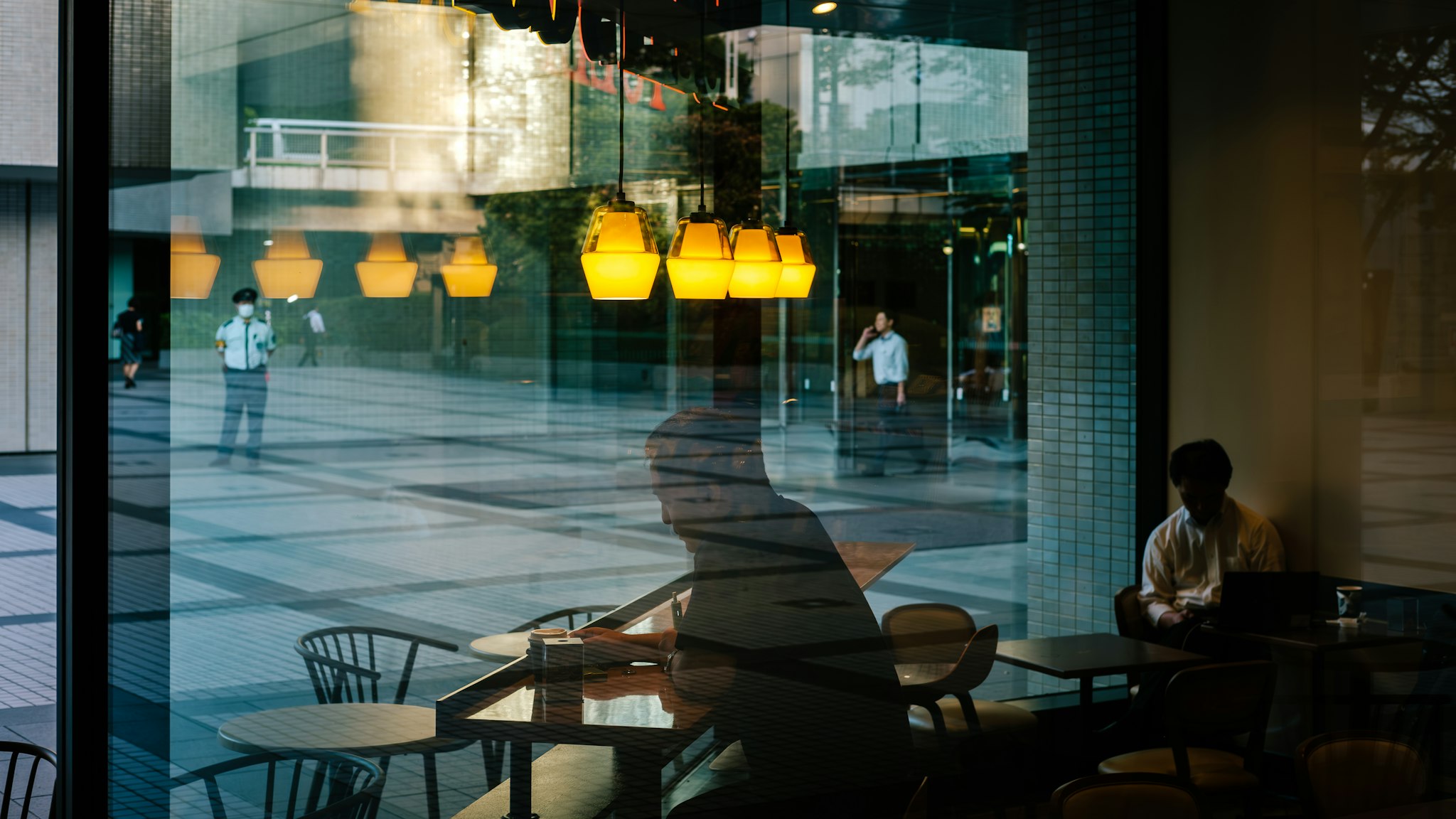 A modern café interior is seen through large glass windows, reflecting a group of hanging pendant lights and silhouettes of individuals both inside and outside, creating a layered, urban street scene.