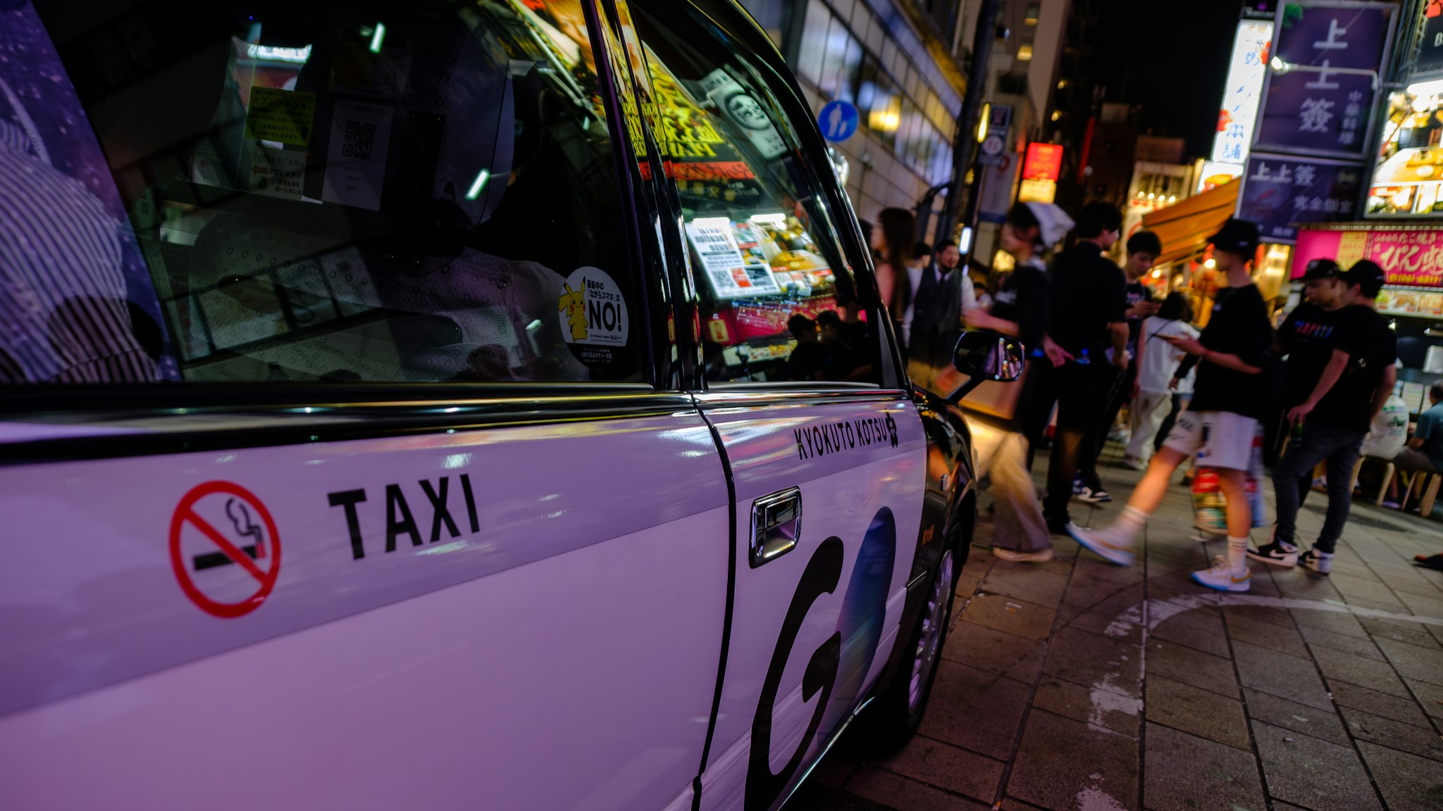 A brightly lit urban street scene at night featuring a white taxi with "No Smoking" signage on the door, parked on a bustling sidewalk filled with people walking by, surrounded by colorful shop signs and illuminated storefronts. SEO keywords: urban night scene, taxi, bustling street.