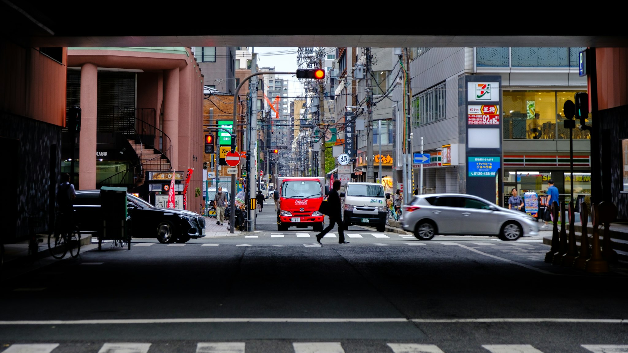 A bustling urban street scene under a bridge features a red delivery truck crossing an intersection, surrounded by cars and pedestrians, with various shops and tall buildings lining the busy road in the background.