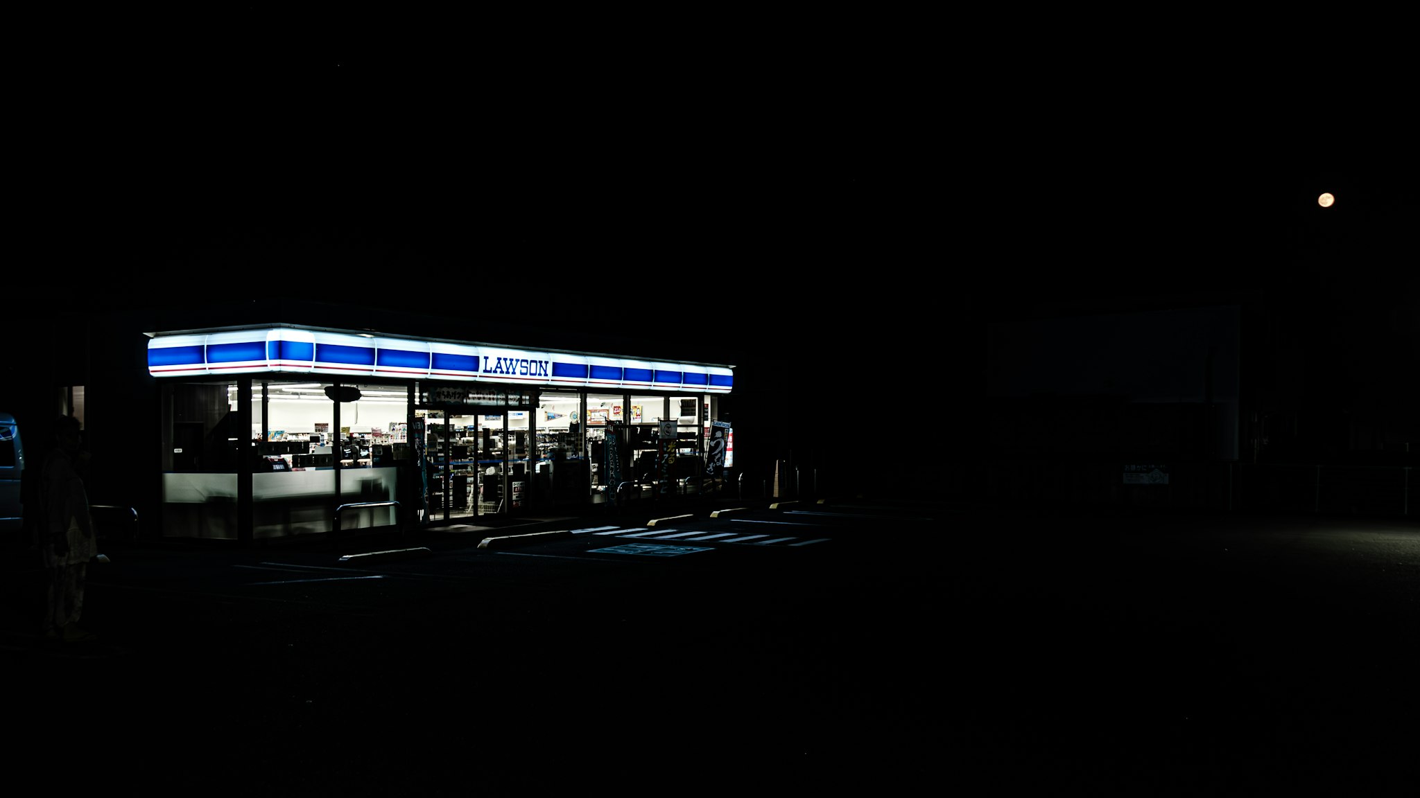 A well-lit convenience store with a blue and white sign stands isolated against a dark night sky, highlighting its bright interior and parking area.