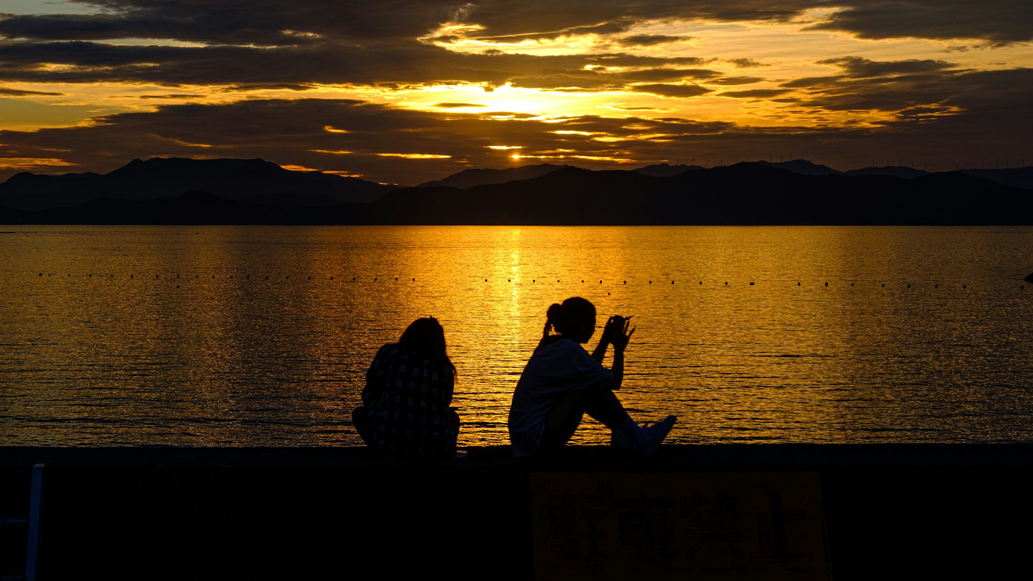 This is a picture of two people sitting on a pier at sunset, enjoying the view of a calm ocean with the setting sun creating a warm orange glow over the water.