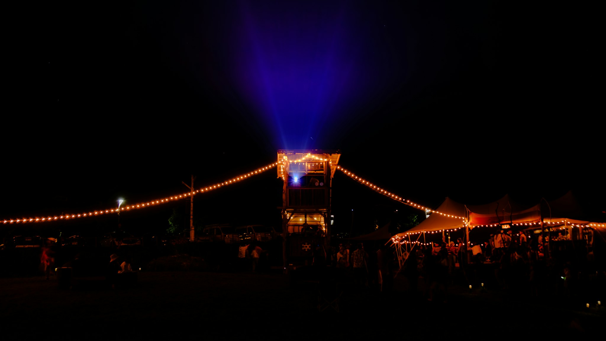 A lively outdoor event at night features strings of warm, glowing lights illuminating a wooden tower and nearby tents, with a vivid blue light beam extending into the sky.