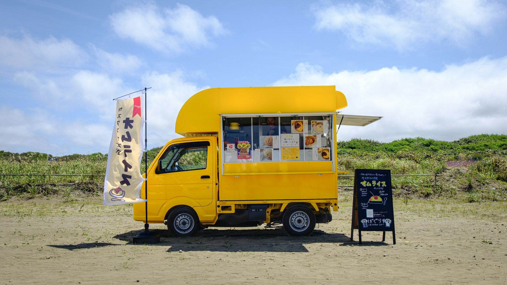 Un vivace furgoncino alimentare giallo è parcheggiato su una spiaggia sabbiosa sotto un cielo parzialmente nuvoloso, con un menu esposto e una bandiera con caratteri giapponesi che ondeggia dolcemente al vento.
